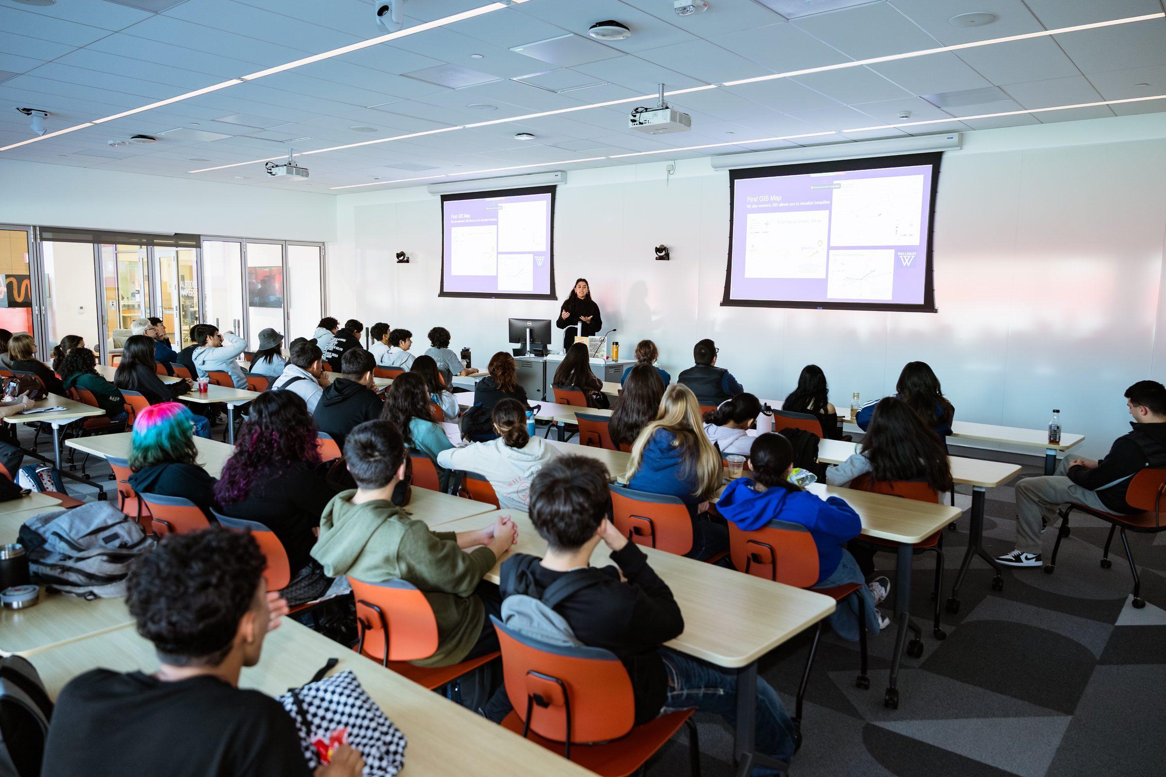 A presenter speaks to a full classroom of students seated at desks, with slides projected on two screens behind her.