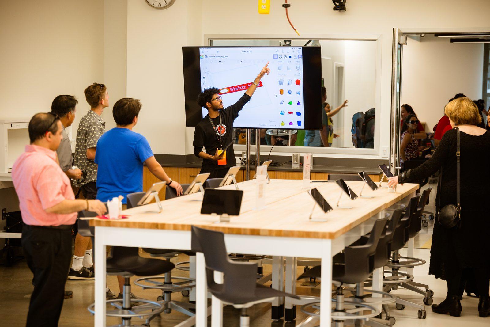 An instructor points at a large screen while students work at tablets in a classroom.